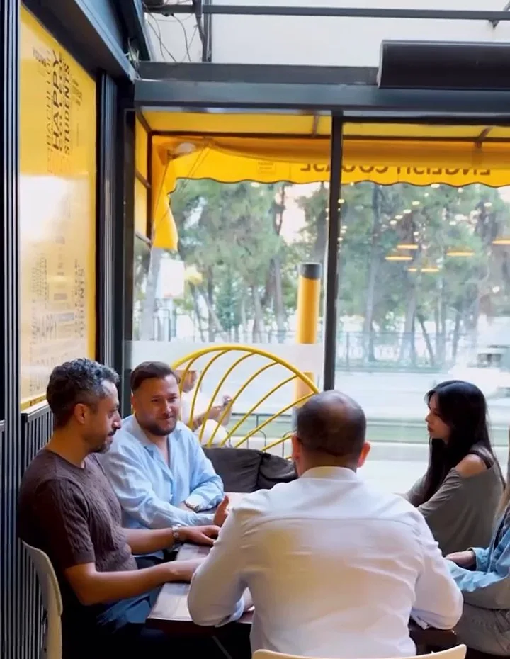 Cam cepheli kafede yetişkinler bir masanın etrafında sohbet ediyor